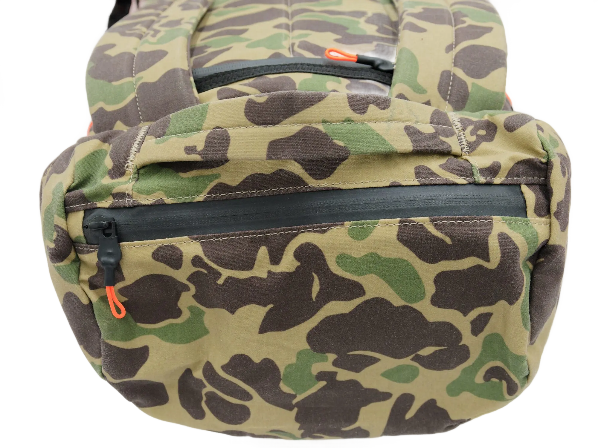 Top-down close-up of the camo backpack showing the horizontal zippered pocket with orange zipper pulls