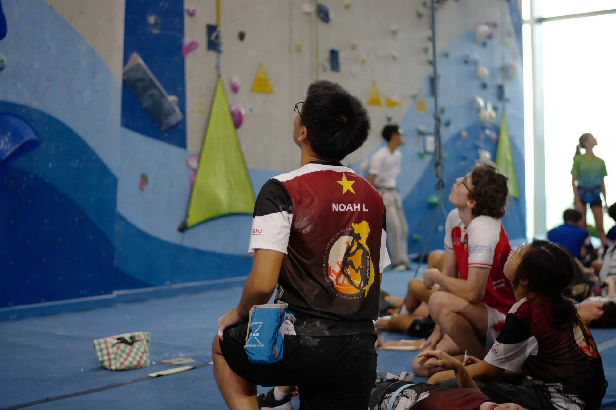 Young climber in burgundy vest waiting during indoor climbing competition at gym wall