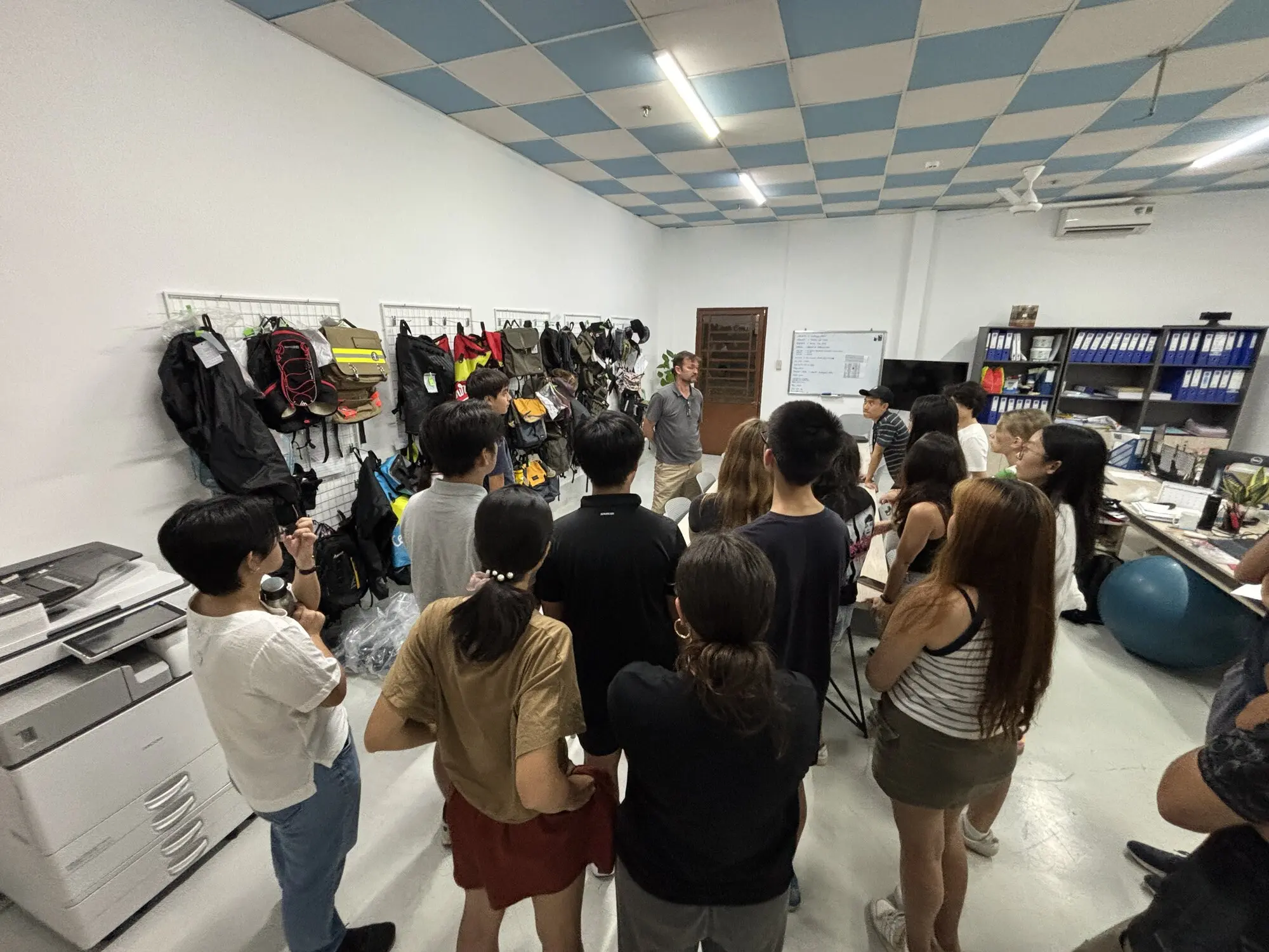 FFG Bags employee presenting to 11th-grade students in factory facility with MOLLE backpacks and tactical bags displayed on wall