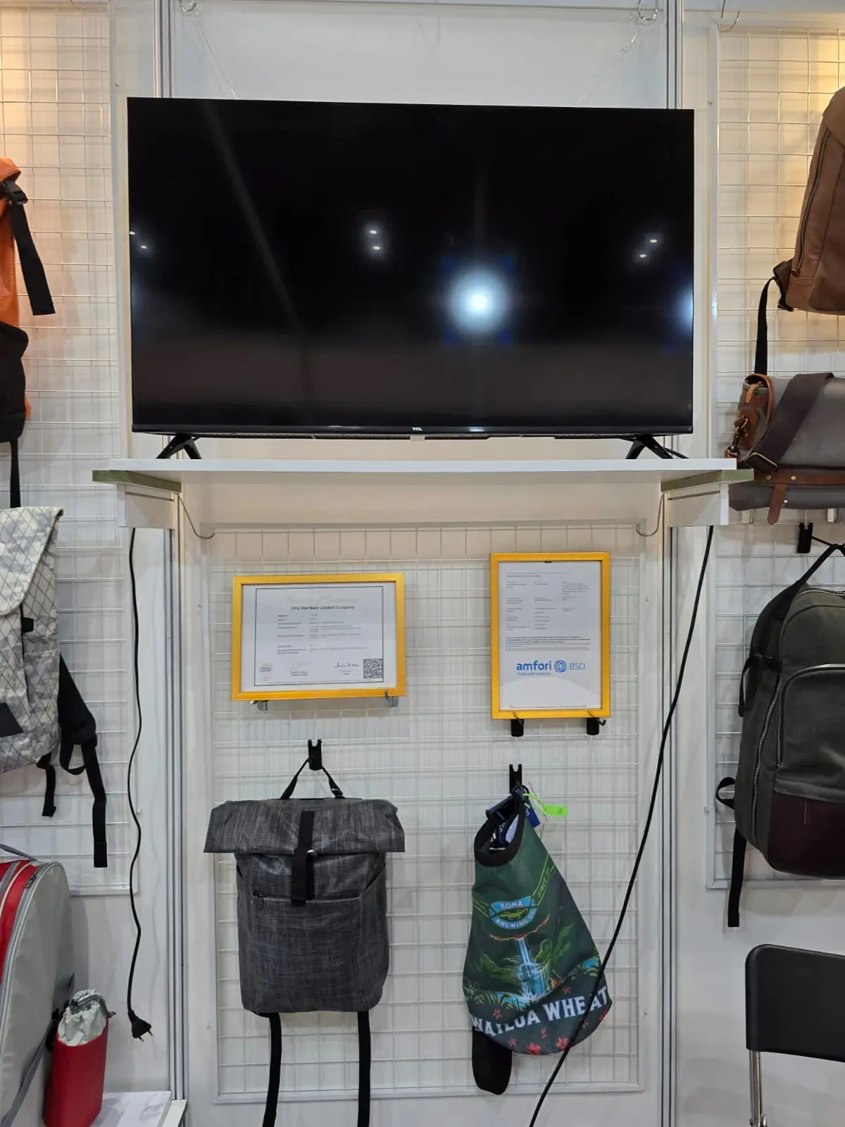 Booth section with mounted display TV, two yellow-framed amfori BSCI certifications, brown leather and tactical bags