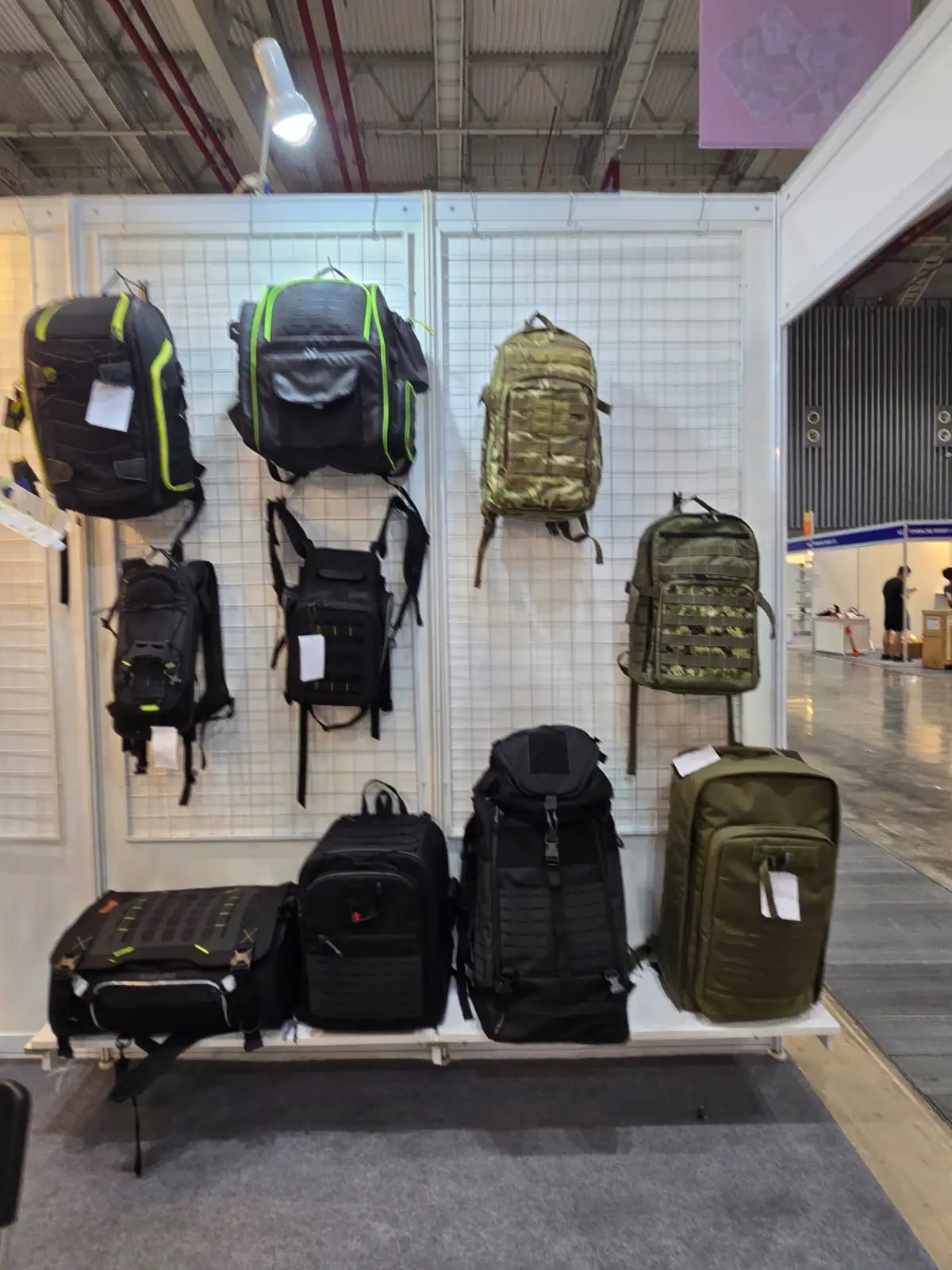 White grid wall exhibiting nine tactical backpacks in black and olive colorways, hung and floor-stacked